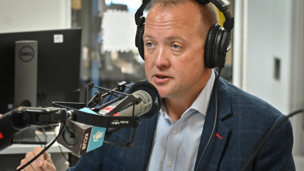 A man wearing a blue sports coat  is wearing a set of headphones, sitting inside a radio studio. He's talking and gesturing with his right hand.