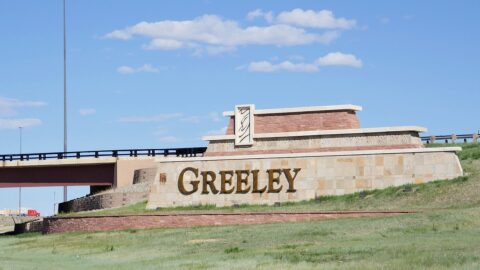 A large brick-and-concrete sign sits in front of a highway bridge. It has a stylized cowboy hat logo and the name "Greeley" on it.