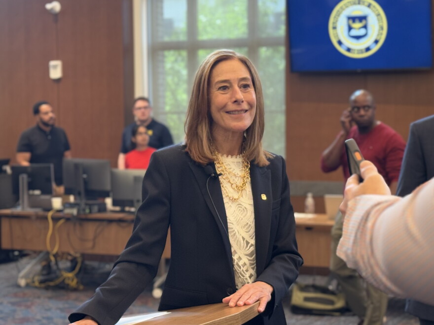 C. Cybele Raver, wearing a navy blazer and white lace top with a gold chain necklace, stands smiling at a wooden lectern during a media interview. In the background, a monitor displays the seal of the University of Michigan.
