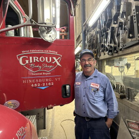 Smiling man in blue workshirt standing by big red red truck which on the door reads 'Giroux Body Shop, Hinesburg, Vermont' 