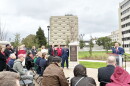 A man in a suit speaks at a podium during a dedication ceremony for a memorial plaque outdoors, with a seated audience watching.