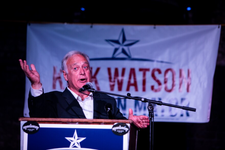 A person standing at a podium gestures as he speaks into a microphone.