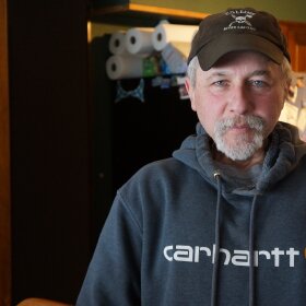 Man with gray beard wearing sweatshirt and baseball cap, standing in kitchen, looking into camera with serious but calm expression