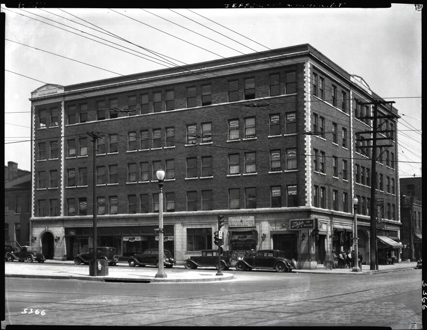 The Peoples Finance Building in Mill Creek Valley was among the buildings demolished in 1959. The black and white image is dated 1935, and photographed from a distance to show the brick, five-story building and five cars parked outside.  A group of people can be seen gathered on the sidewalk. 