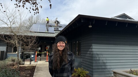A woman stands smiling in front of a building with solar panels being installed on the roof.