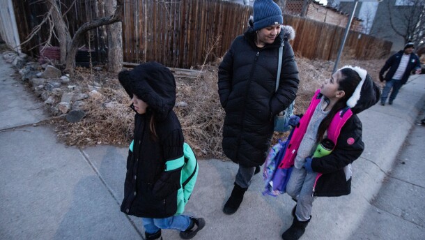 A young girl walks beside her mother on a sidewalk talking with another young girl walking in front of them. All are wearing winter coats and hats.