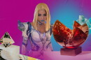 A cosplayer wearing a blonde wig and fake white and blue armor and blue lipstick. There is glass art displayed on both sides of her, on top of a hot pink background.