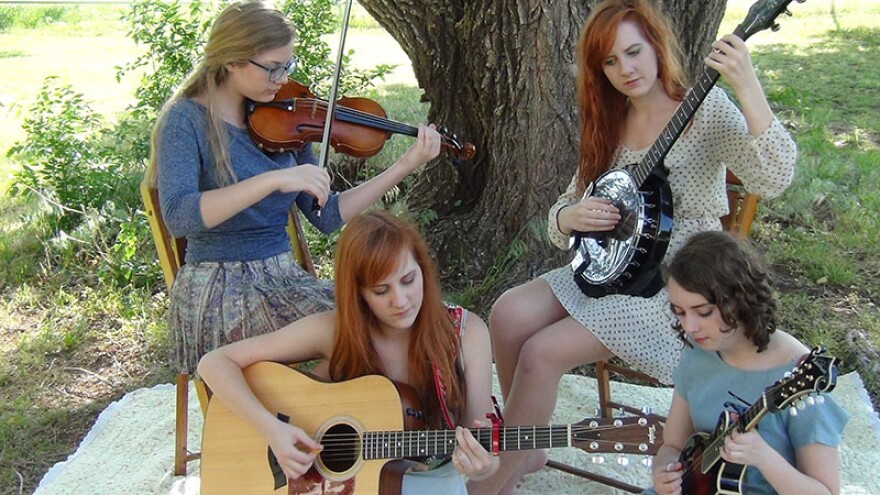 4 young musicians seated on a picnic blanket by a tree, playing fiddle, banjo, guitar and mandolin