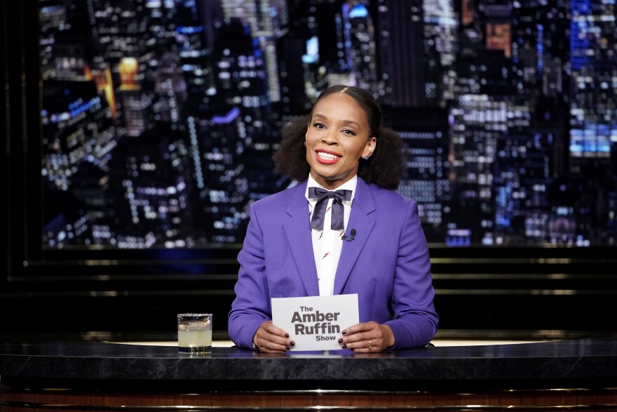 Amber Ruffin sits behind a desk wearing a purple suit and black bowtie