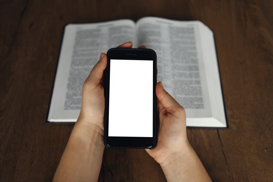 A woman's hands hold and a smart phone touch with a blurred Bible behind her hands