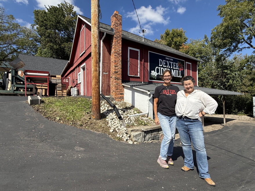 two women stand on a black driveway in front of a red barn that has a "Dexter Cider Mill" sign on it