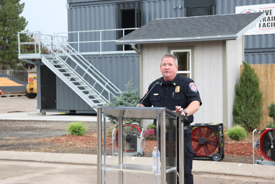 A man in a fire chief uniform with grey hair stands at a clear plastic podium, speaking into a microphone. Behind him is a small mock house, and behind that are large grey shipping containers with a metal staircase leading up to them.