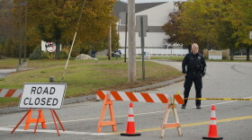 A police officer stands at a road closure near a bowling alley, seen in background, Thursday, Oct. 26, 2023, in Lewiston, Maine. The site is one of Wednesday's two mass shootings in the city. Residents have been ordered to shelter in place as police continue to search for the suspect.