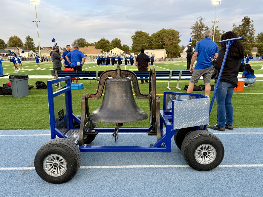 A big iron school bell, mounted to a metal cart, sits on the blue track of Walla Walla High School's Blue Devil Stadium while cheerleaders, band students and others wait on the football field for the game to begin.