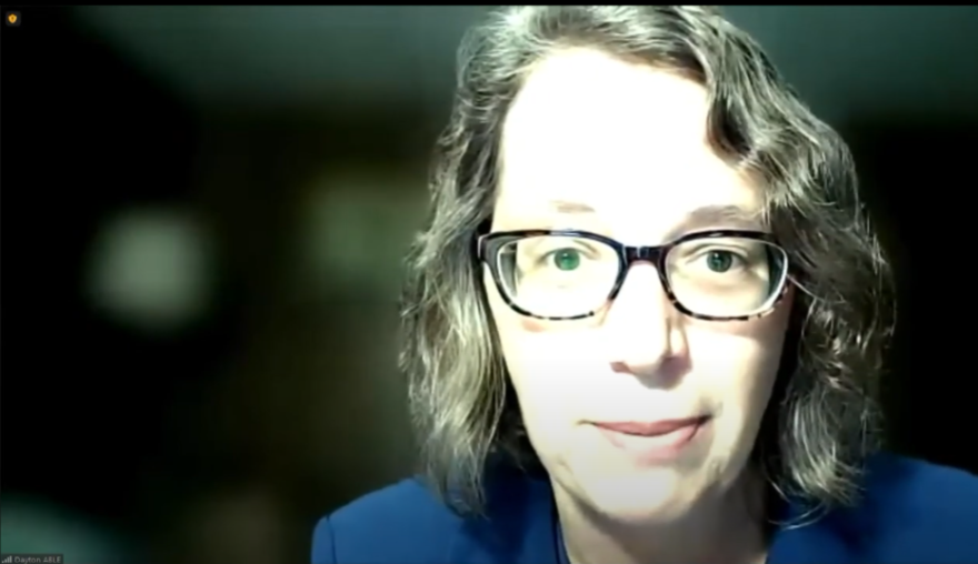 An up-close screen capture of Debra Lavey. She has shoulder-length blonde hair, a blue jacket and glasses.