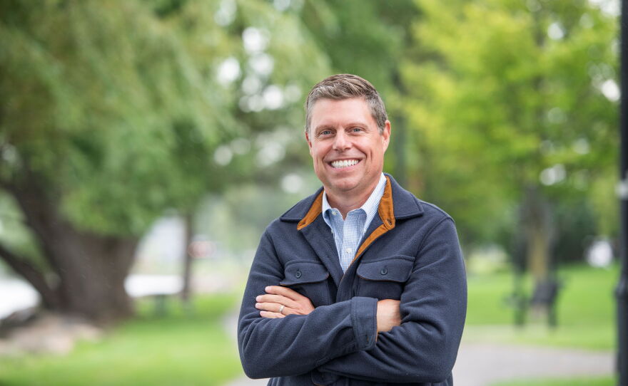 A smiling man with short brown hair standing with his arms crossed. He is wearing a blue collared button-down shirt under a blue fleece with a brown lining. 