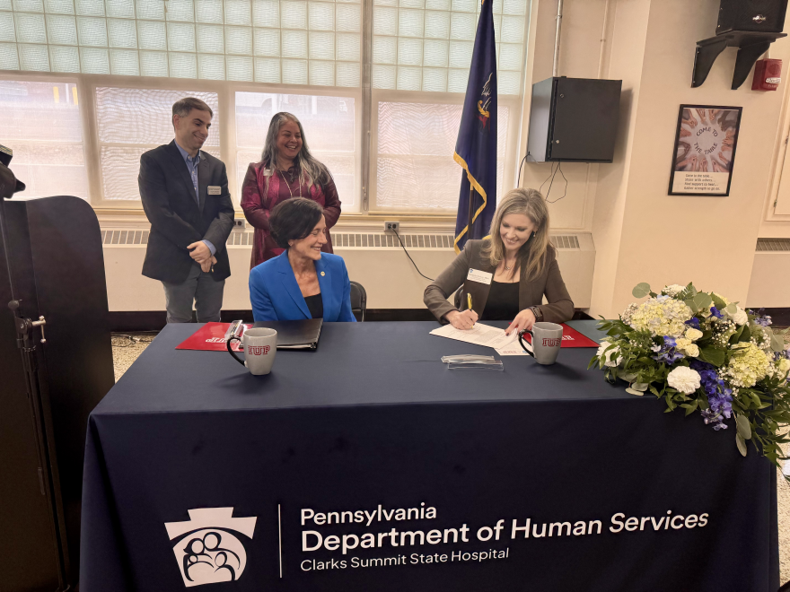 Kimberly Tenbus, CEO of Clarks Summit State Hospital (right), signs the clinical agreement while Pa. Human Services Sec. Val Arkoosh (left, seated), Dr. Ryan Smith, the associate dean of clinical affairs and graduate medical education at IUPCOM (standing left) and IUPCOM's founding dean Dr. Miko Rose watch.