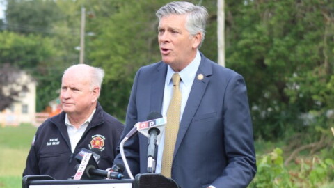 Darin LaHood speaks at the Bloomington Parks & Recreation podium as Mayor Dan Brady stands behind him in his fire department jacket.