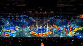 Three rings of a circus shine bright with performers, trapeze artists, and bright lights.