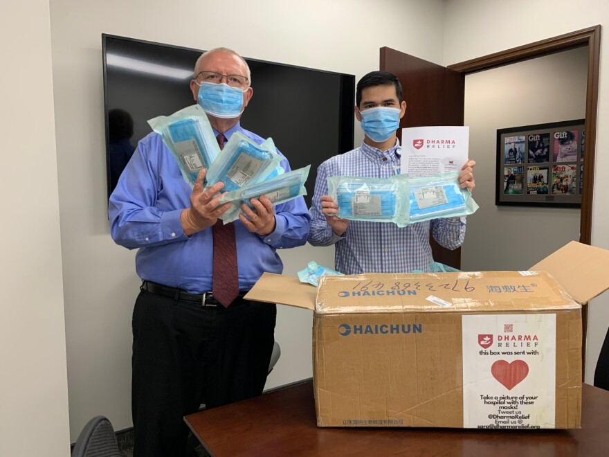 Two men wearing surgical masks hold up packages containing more surgical masks. They are in what appears to be an office. A box is on the desk. It is labeled "Dharma Relief."