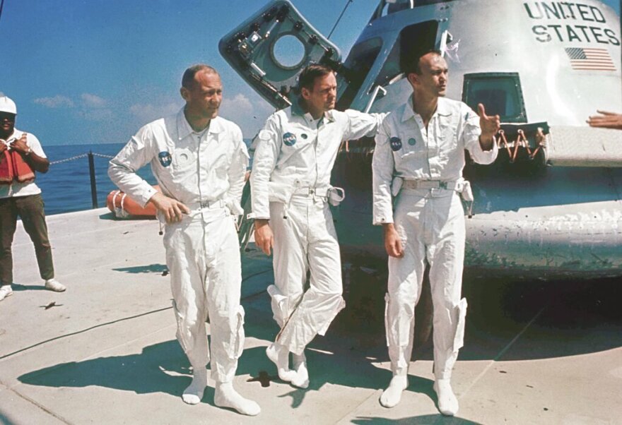 Three men in white jumpsuits stand in front of a spacecraft. They appear to be talking with someone out of frame to their left.