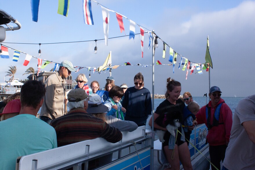 Flags strung around the top of the boat move in a slight breeze while many people stand and prepare to step off the boat. A woman holding a toddler under one arm and a dog under the other is stepping from the boat onto the dock while a man in a life vest holds open the boat's gate.