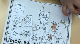 A child's hand can be seen on a table with a worksheet that is intended for coloring and learning. 