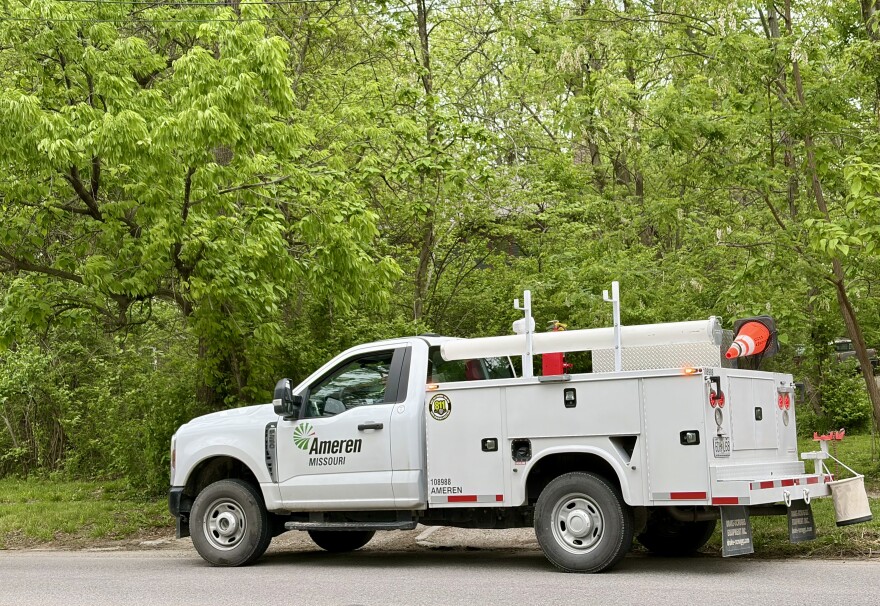 An Ameren Missouri utility truck is parked on a residential city street in Columbia Missouri. Behind the truck the frame is filled with lush green trees. 