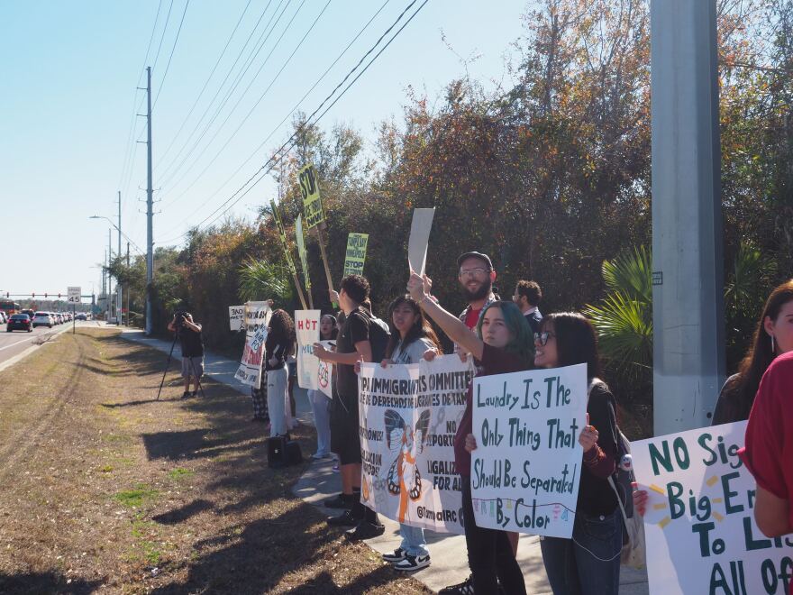 Students and community organizers stand on the sidewalk with signs condemning immigration enforcement.