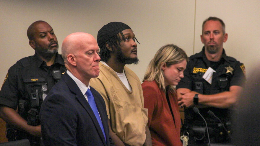A prisoner in a court looks up. He stands between two lawyers who look disappointed. On either side of them, Sheriff's deputies look on. 
