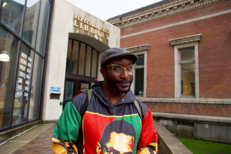 A man in gold-rimmed glasses and a Malcolm X shirt stands outside a library.