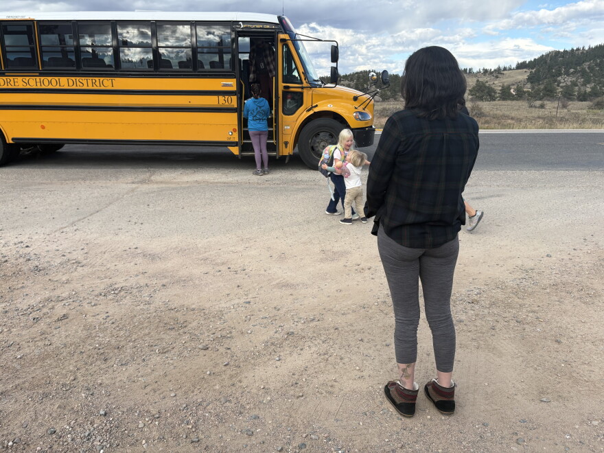 A woman wearing a dark shirt waits in front of a yellow school bus.