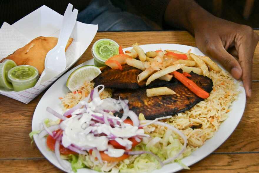 An oval white plate sits on a table. A man's hand is touching the side. The plate shows a salad with white dressing, rice and blackened fish. Beside the plate is a cardboard container with a brown, fried pastry.