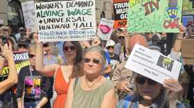 Crowd of people holding signs, workers demand living wage and health care for all and Hey billionaire cheapskate, our workers deserve a living wage. Pay up. 