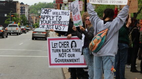 Demonstrators stand on a sidewalk holding signs that read "Let Cher turn back time, not our government!" and "GOP will this apply to your mistresses or just us?"