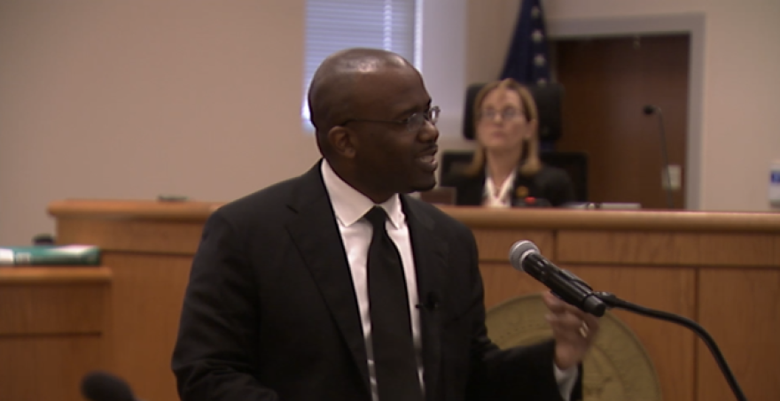 A man in a black suit and tie speaks into a microphone in a courtroom, with a judge seated in the background.