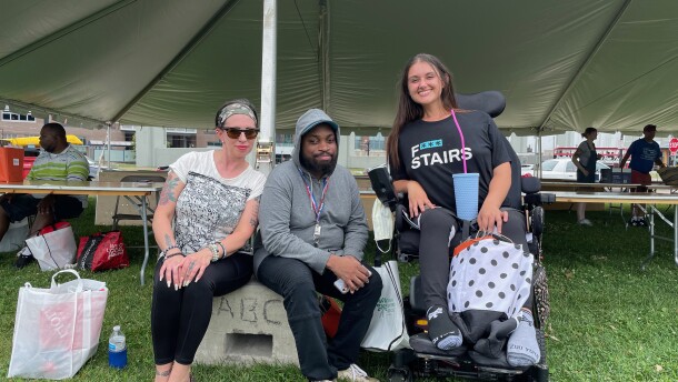 Three people pose together for a photo in front of a large event tent. One person is using a motorized wheelchair and the other two people are sitting on a concrete block.