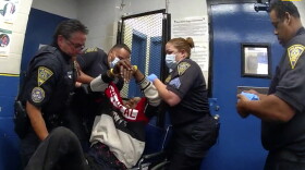 A screenshot from New Haven Police Department body camera footage shows a grimacing Richard Cox being lifted into a wheelchair by NHPD officers. Cox, 36, was severely injured and likely paralyzed moments before while being en route to NHPD headquarters in a transport van. Cox's head slammed into the front interior wall of the van after the driver braked suddenly. Cox is in the hospital and several NHPD officers are on paid leave while the incident is being investigated.