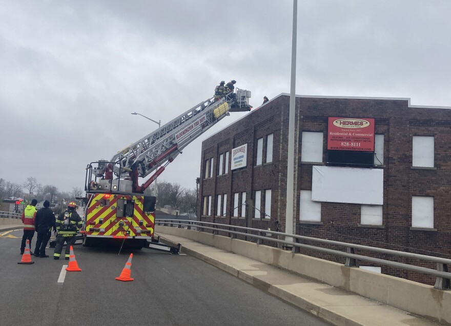 A fire department ladder truck on a bridge with ladder extended to the top of a warehouse building on the right. Firefighters stand to the left of the vehicle.