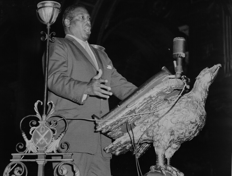 A man in a suit speaks passionately at a podium shaped like an eagle, with a microphone in front of him. Ornate metalwork and a decorative lamp are visible in the foreground. The background is dark and indistinct.