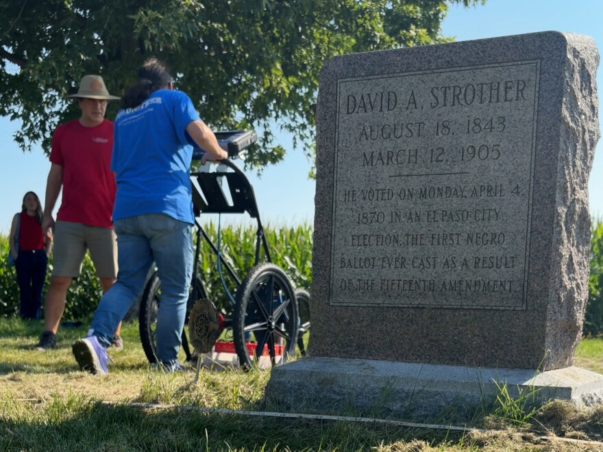 a memorial gravestone is in the foreground. In the background, a volunteering pushes an underground radar device while a man in a red t shirt watches