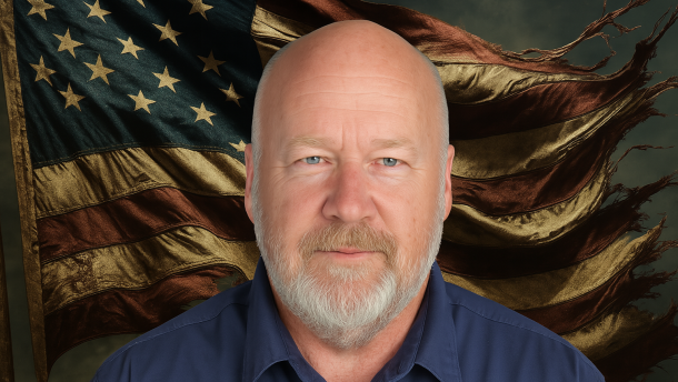 A man wearing a dark blue collared button-up shirt looks into the camera. A tattered American flag waves in the background.