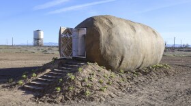 The outside of the Idaho Potato AirBnB. There are stairs leading up to the giant tuber and the door is open. The potato sits on top of a mound of dirt and it is surrounded by dirt as well. 