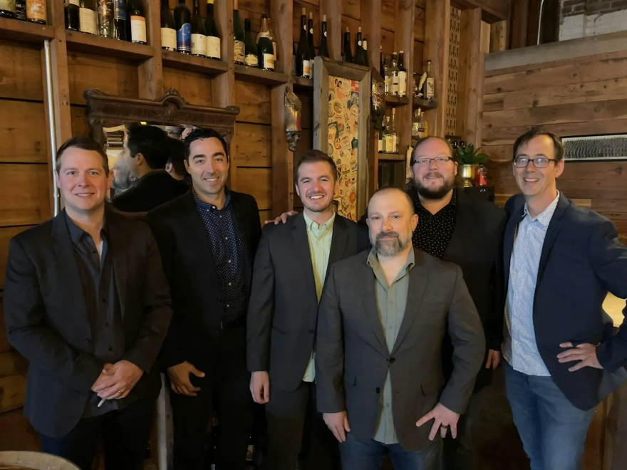 Six musicians are standing together as a group and smiling without their instruments.