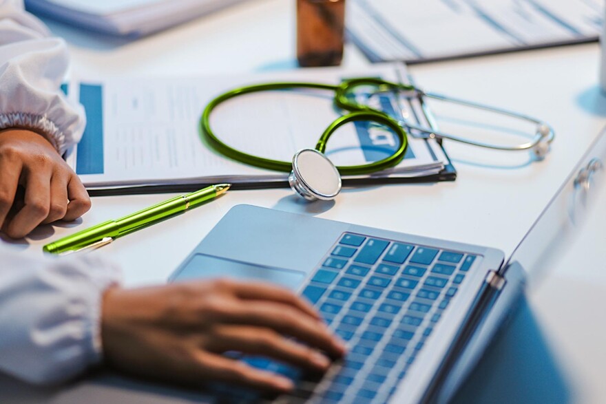 Doctor sitting at a computer with a green pen and stethoscope. 
