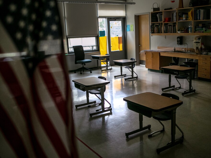 a dimly lit classroom with wooden desks spaced apart from each other, an American flag in the foreground and a door illuminated in natural light toward the back of the classroom.