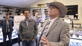 a man in a cowboy hat stands talking and gesturing in a gun store