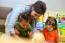 A woman smiles as she interacts with three young children in a classroom, surrounded by colorful books and educational materials.