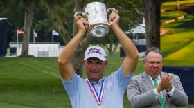 Senior golf player Padraig Harrington lifts up a trophy over his head while a tournament official looks on and claps.