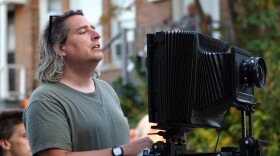 A heavyset man with long grey hair stands behind a camera.